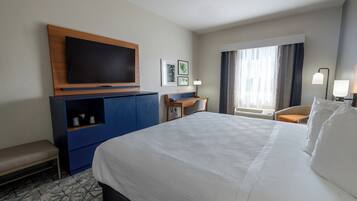 A hotel room with a large bed, a flat-screen TV mounted on a wooden cabinet, a desk with a chair, and a window with curtains.
