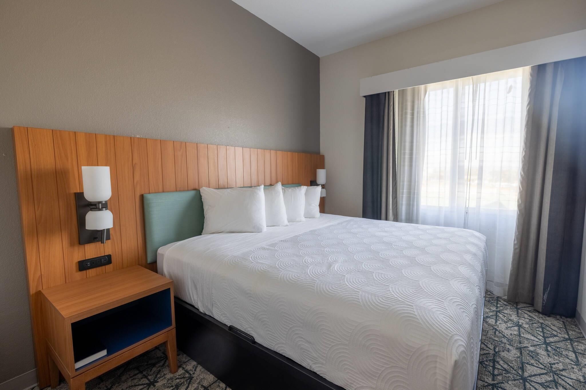 Ein Hotelzimmer mit einem hölzernen Kopfteil, einem Bett mit weißer Bettwäsche, einem Nachttisch mit einer Lampe und einem Fenster mit Vorhängen.