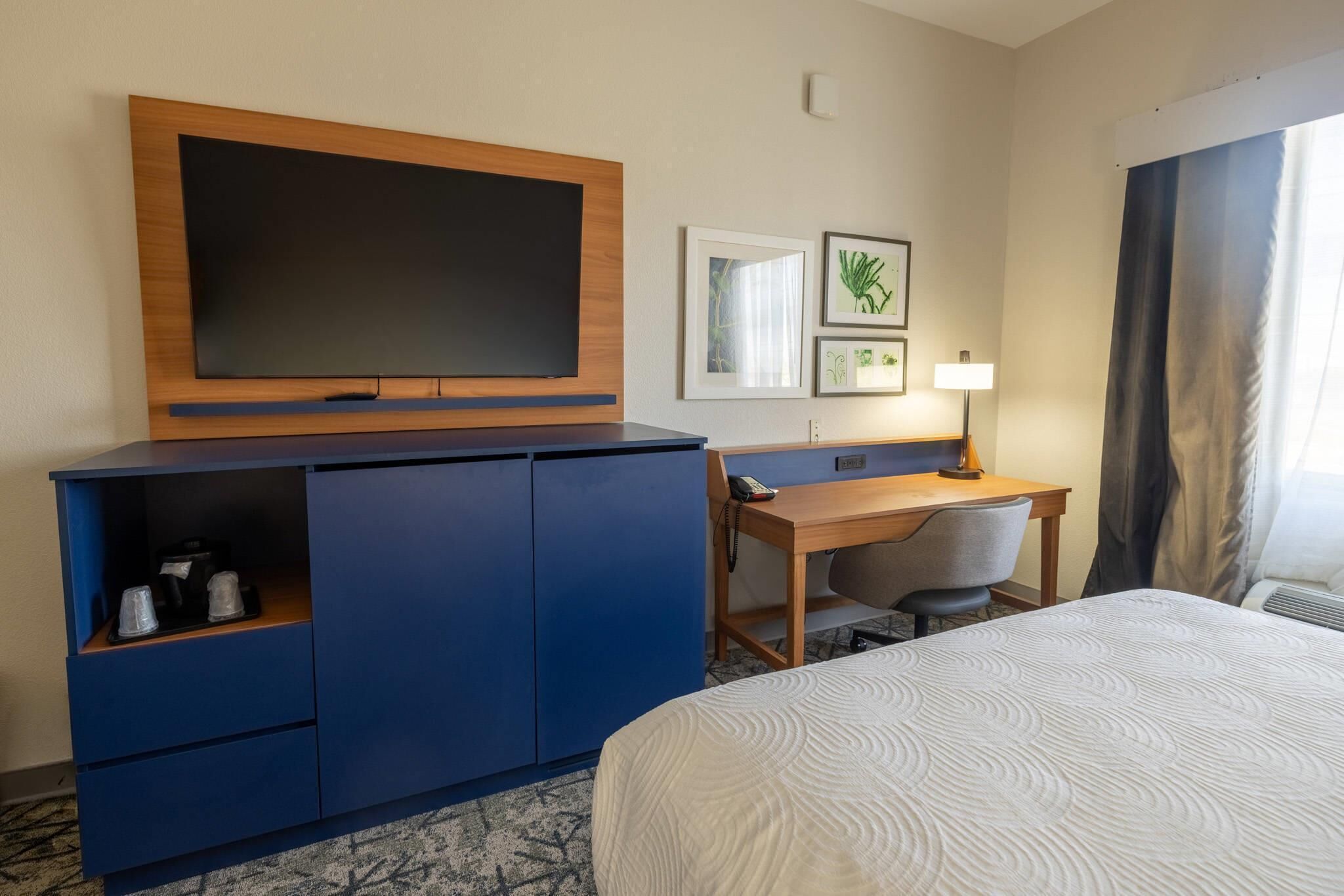 Habitación de hotel con cama, escritorio, silla, televisor, cafetera y lámpara.