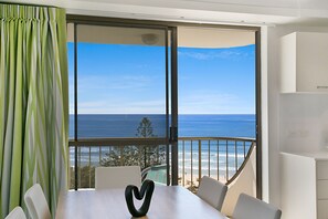 Habitación con un amplio ventanal corredero que da a un balcón con vistas al océano. Se observa una mesa con un adorno en forma de corazón y varias sillas.
