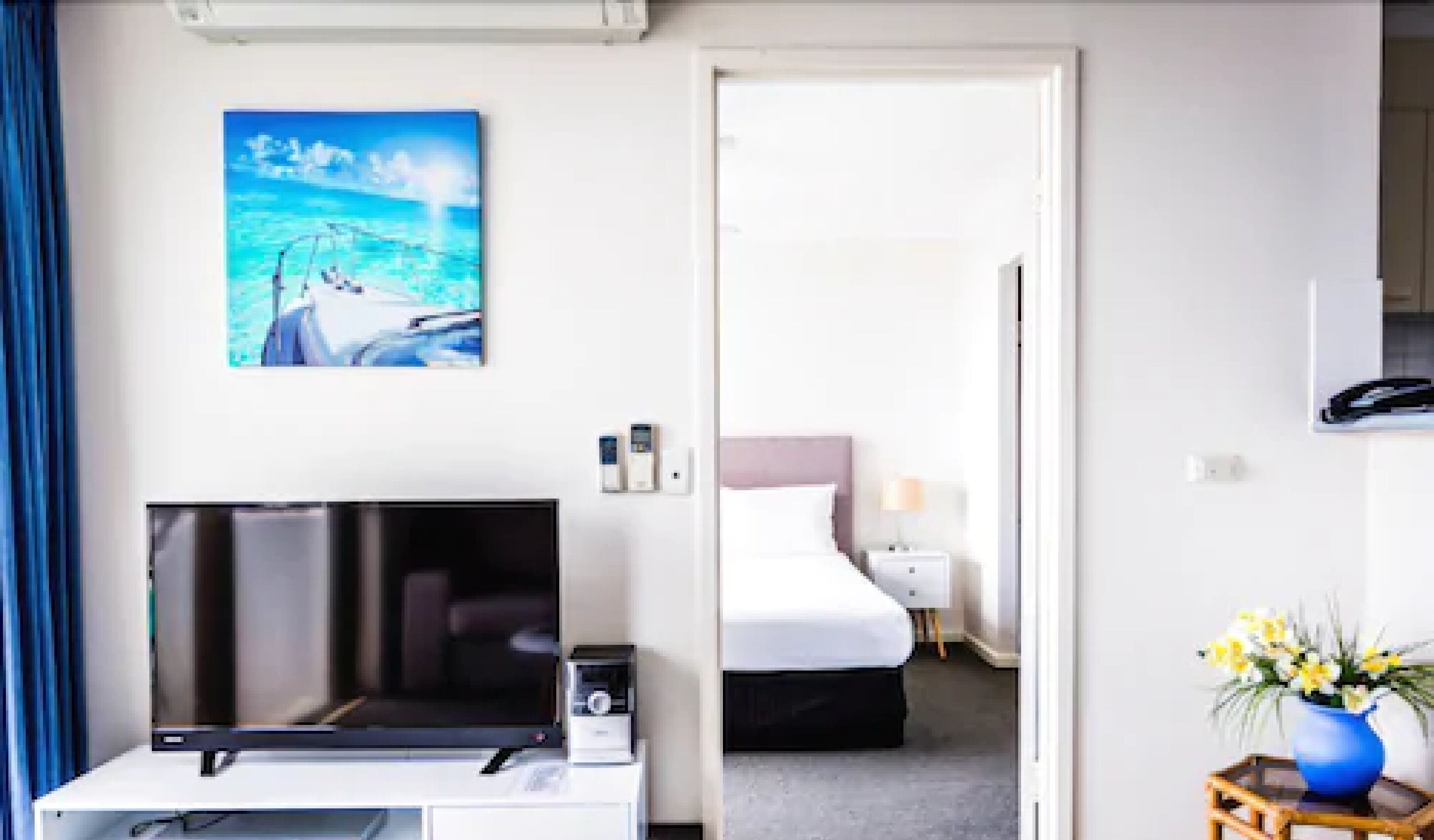 A hotel room with a bed, a TV, a painting, and a vase with flowers.