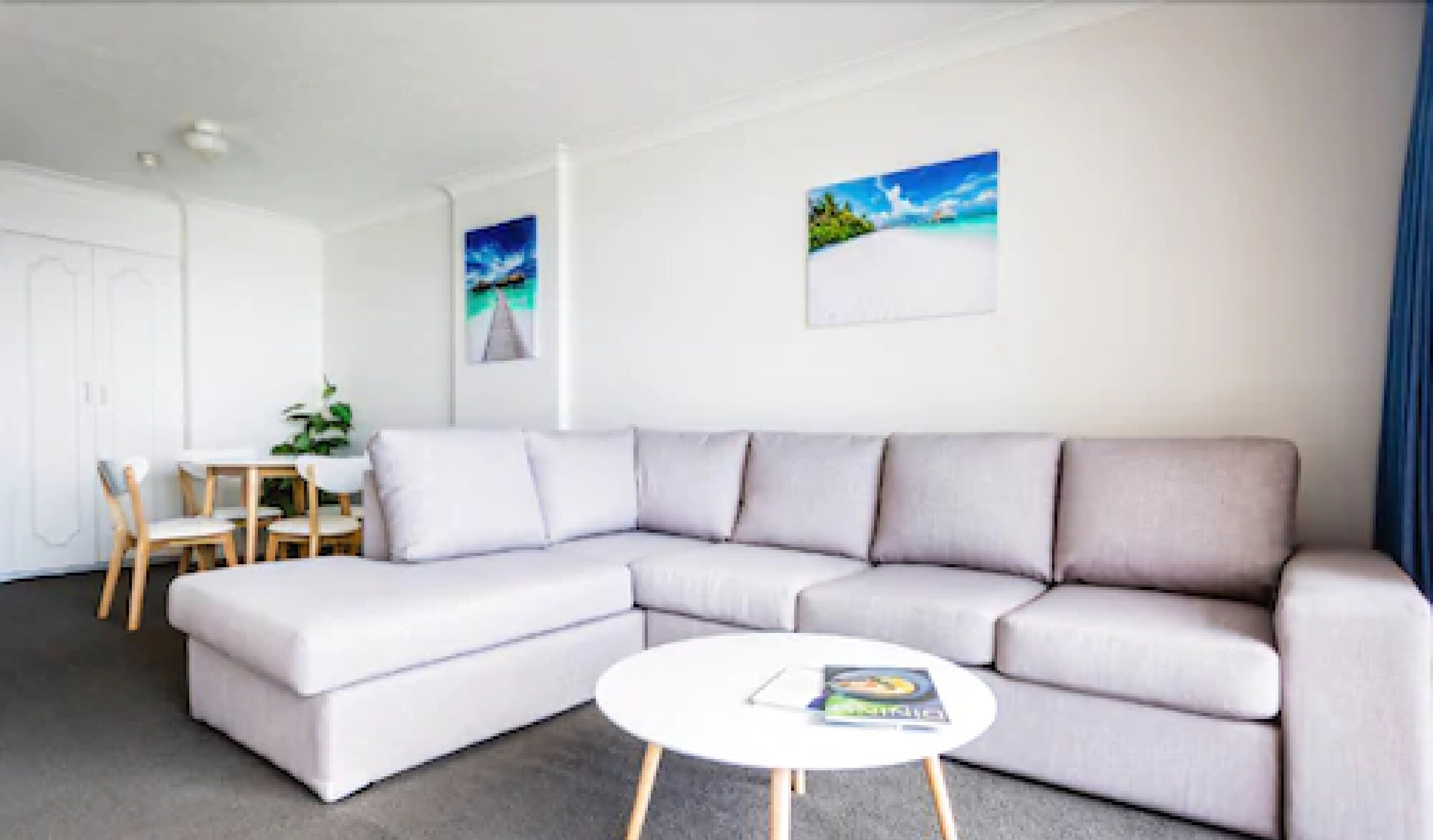 Un salon moderne avec un canapé d’angle gris, une table basse ronde et un coin repas comprenant des chaises en bois et une petite table.