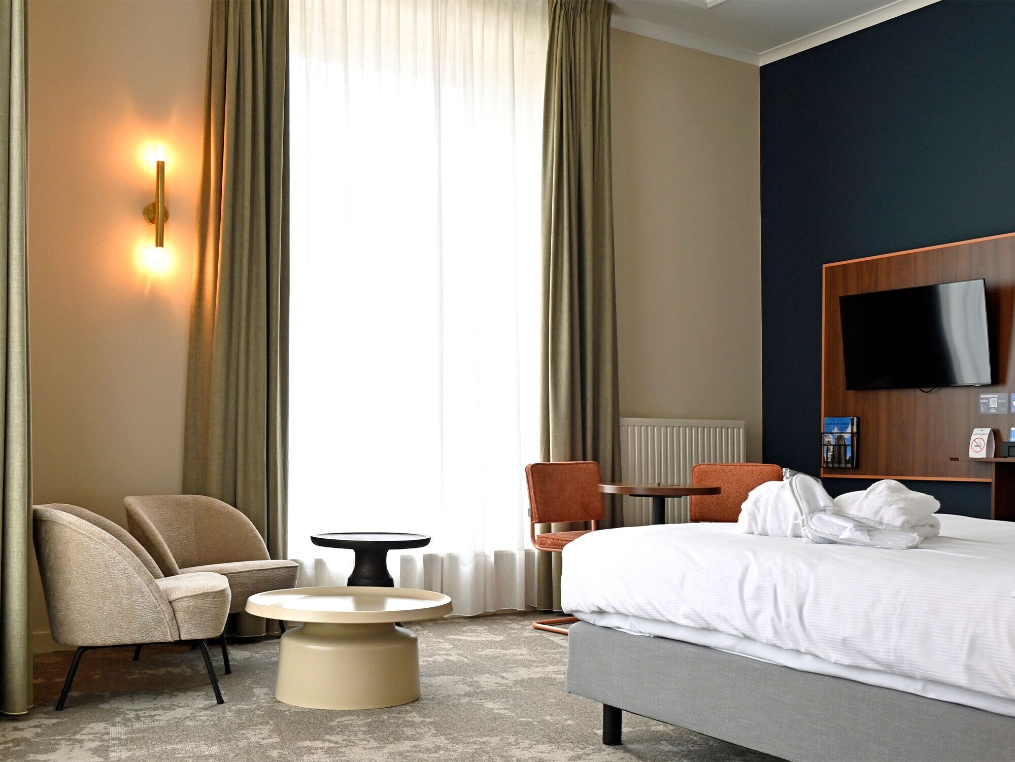 A hotel room with a bed, two armchairs, a small round table, a TV mounted on the wall, and a window with curtains.