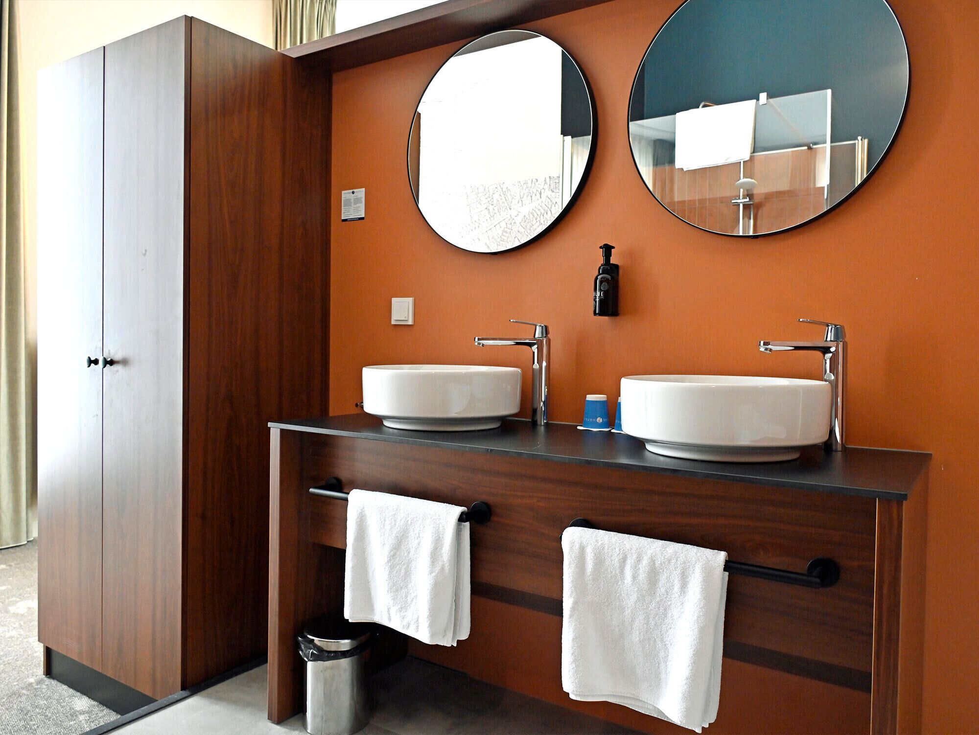 A bathroom with two sinks, a mirror, a towel rack, and a trash can.
