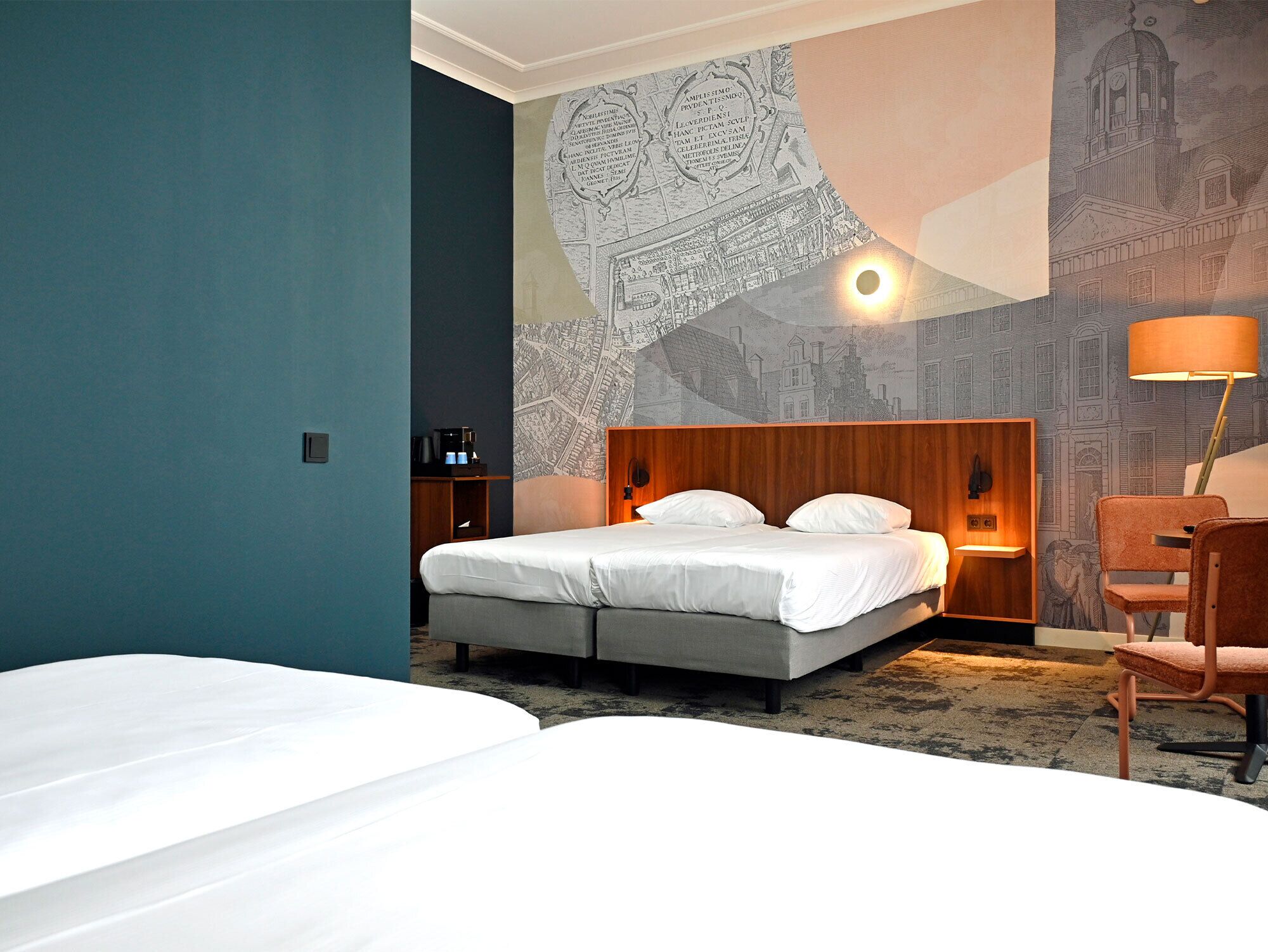 Habitación de hotel con cama, mesita de noche, lámpara y un mural en la pared con vista urbana.