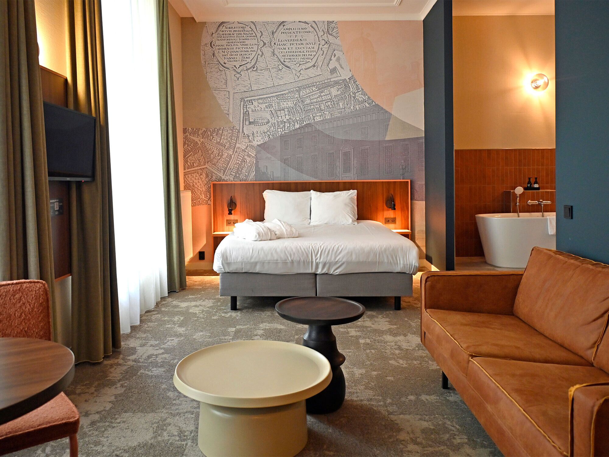 A hotel room with a large bed, a sofa, a round coffee table, a TV, and a bathroom with a bathtub.