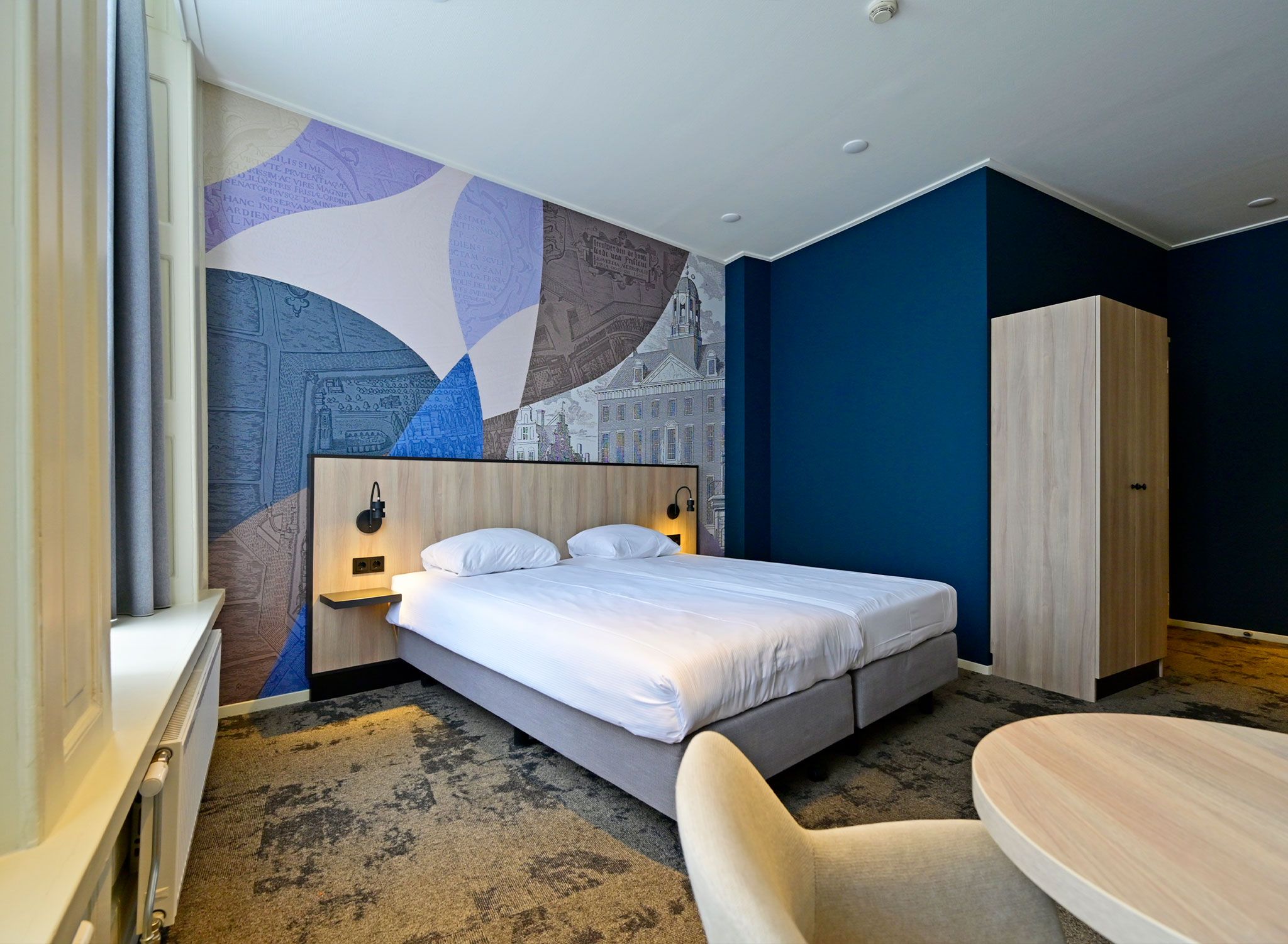Quarto de hotel com uma cama grande, uma escrivaninha, um guarda-roupa e uma parede com um mural da paisagem urbana.