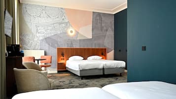 A hotel room with a large bed, a desk with a chair, a wall with a map design, and a nightstand with a lamp.