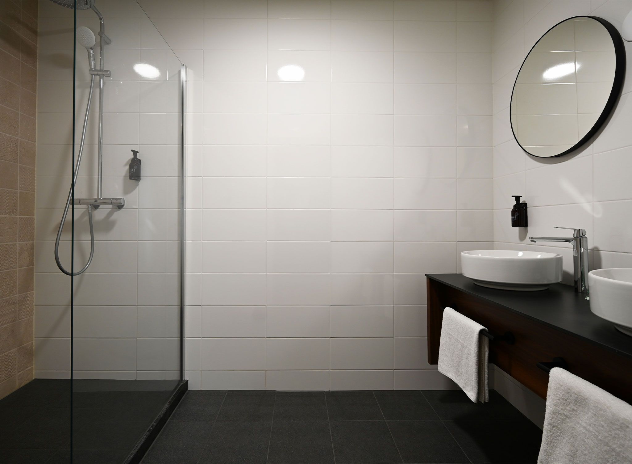 A modern bathroom with a glass shower enclosure, white tiled walls, and a dark countertop with two sinks.
