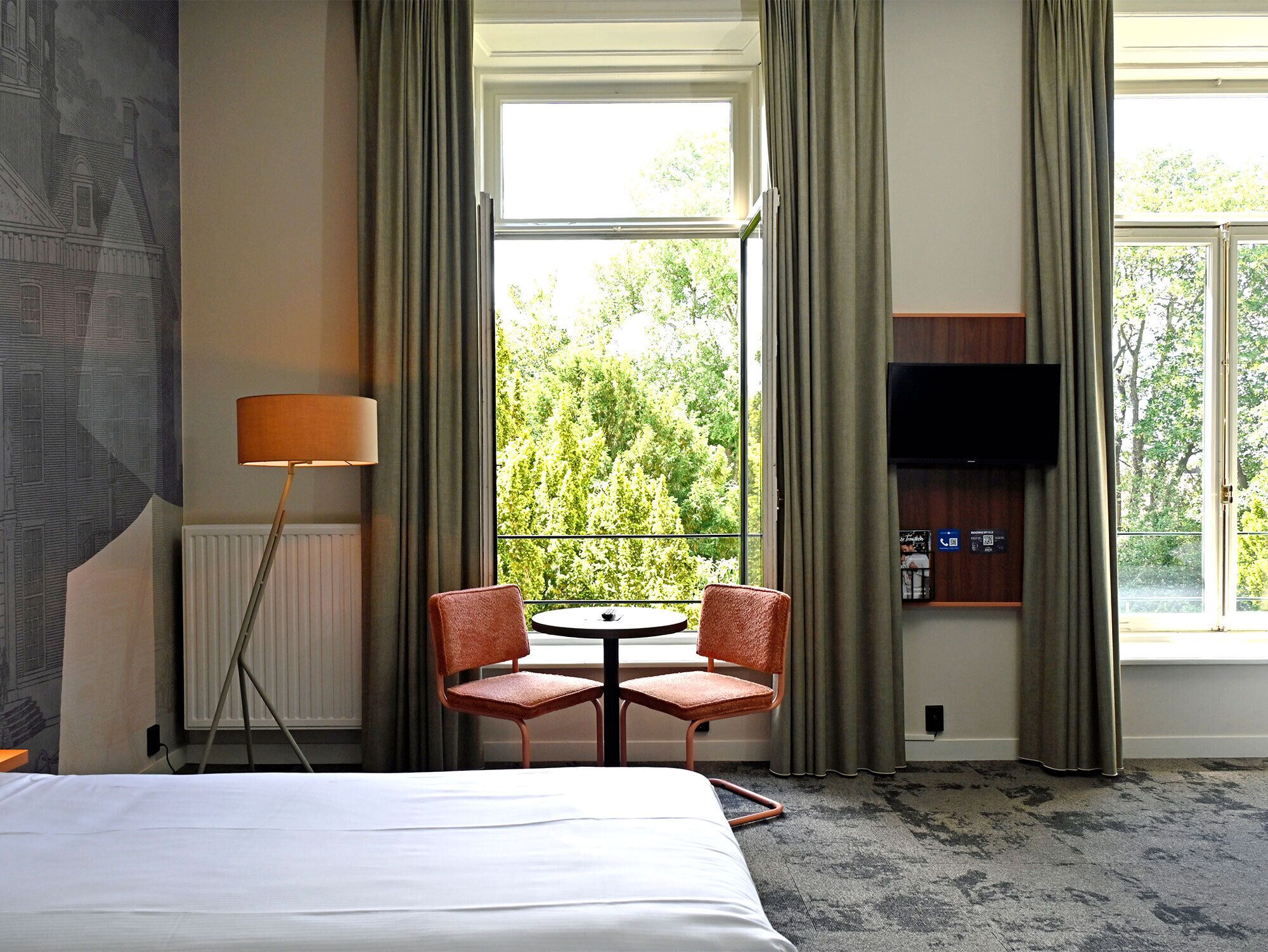 A hotel room with a bed, a small table with two chairs, a lamp, a TV, and a window with curtains.
