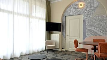 A hotel room with a large window, a flat-screen TV, a round dining table with chairs, and a wall with a detailed map mural.
