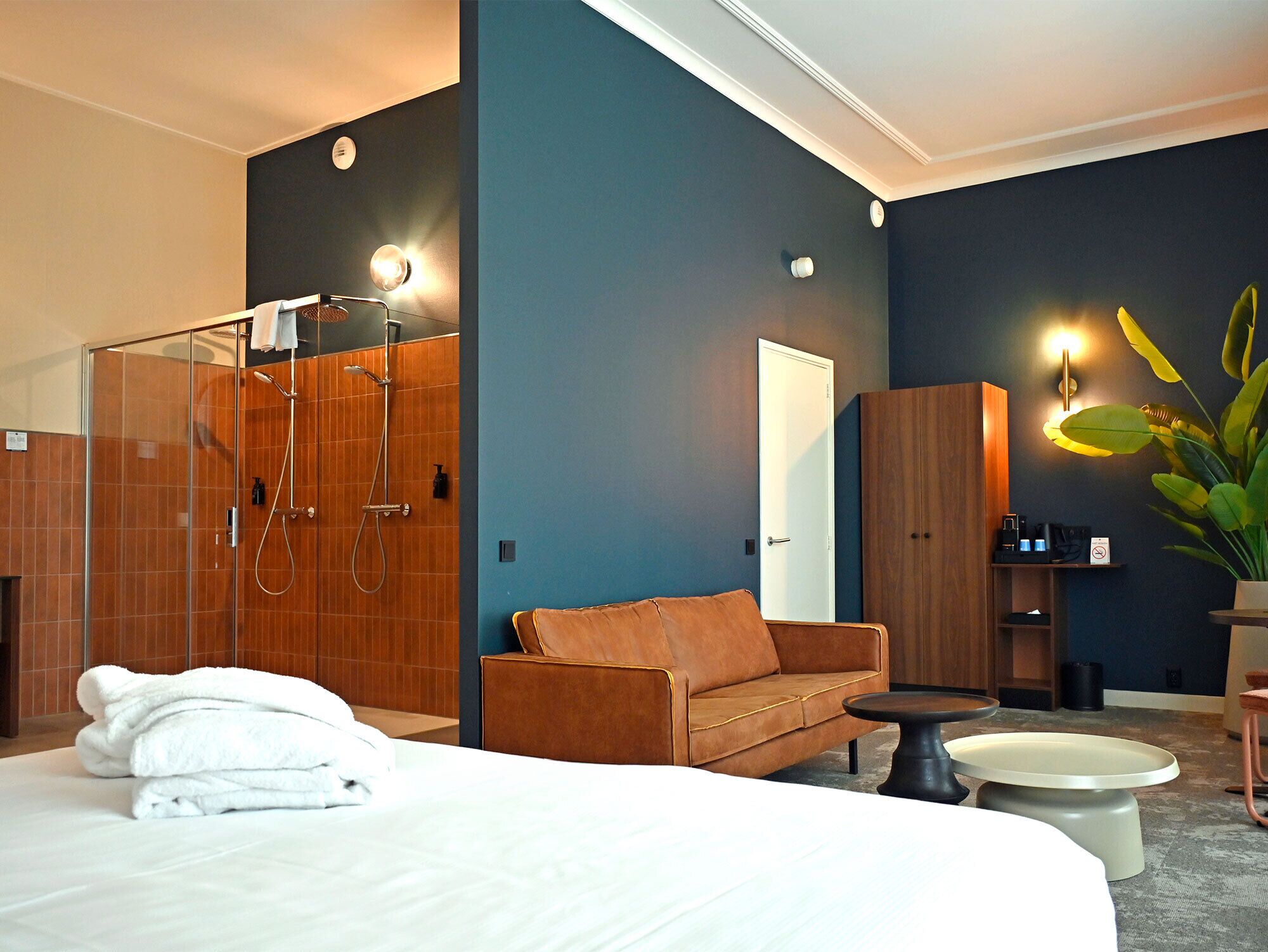 A modern hotel room with a large bed, a shower, a sofa, a small table, and a cabinet.