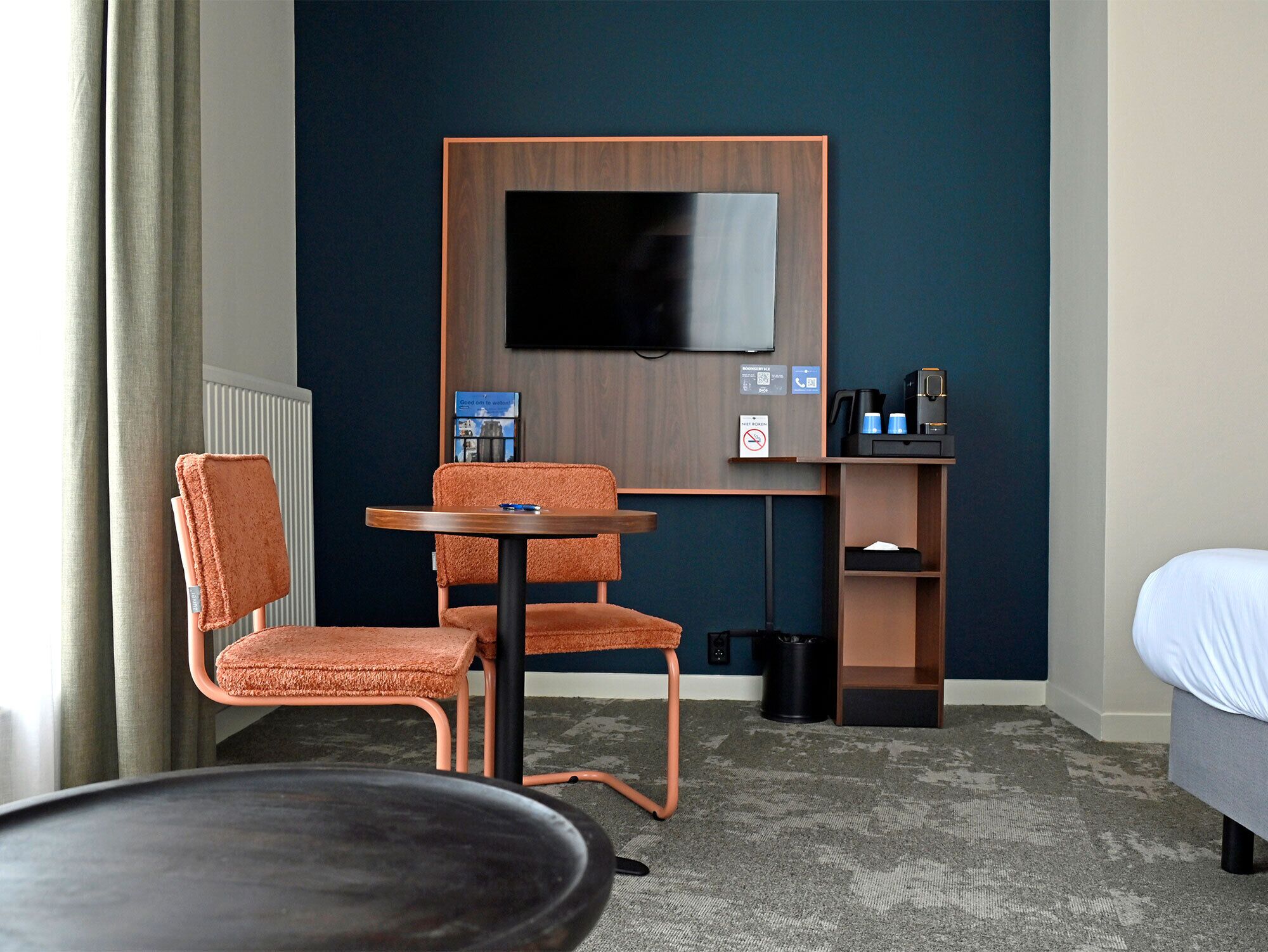 A hotel room with a TV mounted on the wall, a desk with a chair, a bed, and a small table.