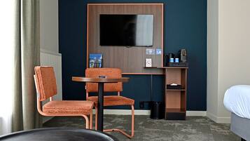 A hotel room with a TV mounted on the wall, a desk with a chair, a bed, and a small table.