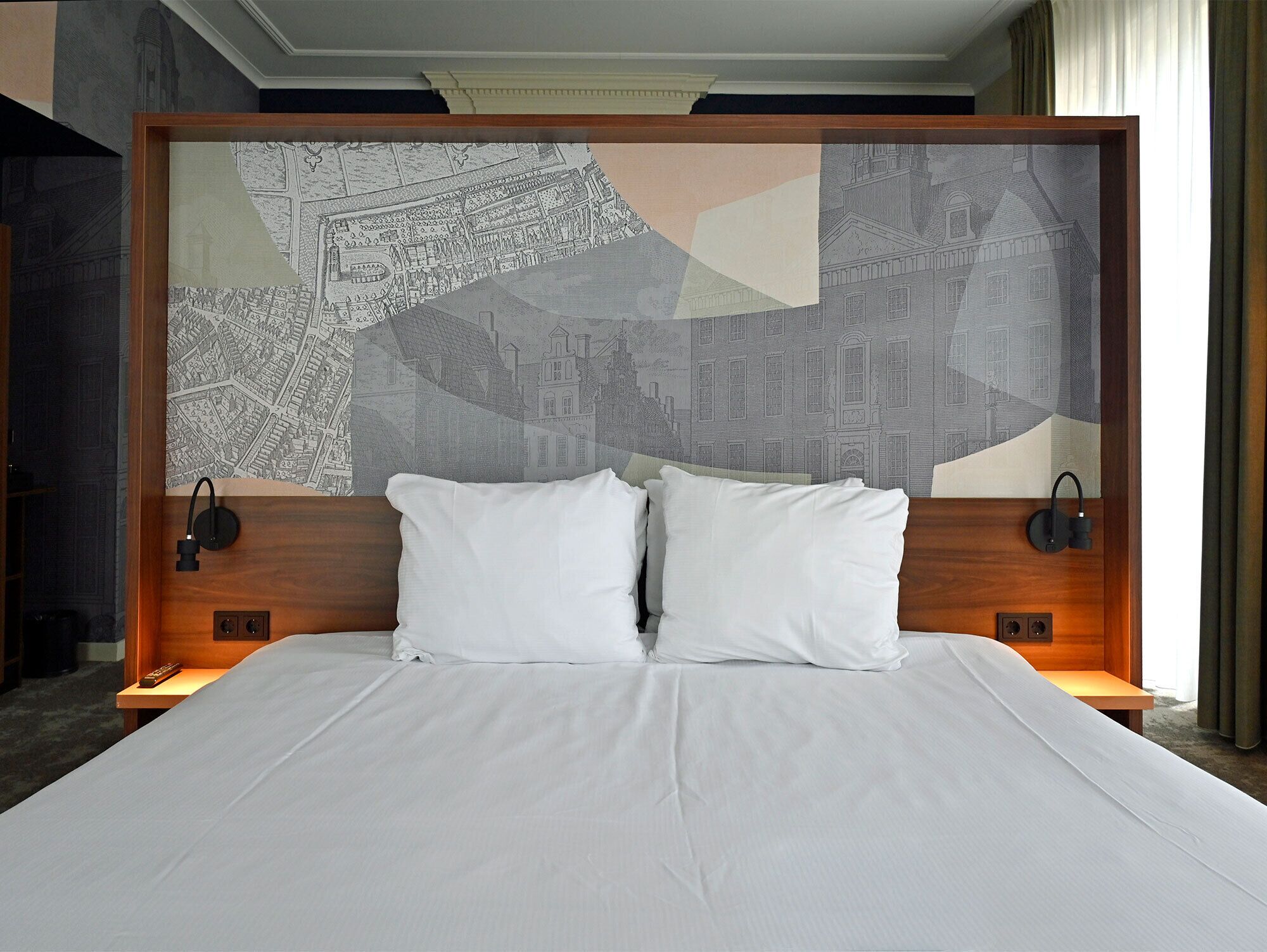 Una camera d'albergo con un letto grande, due cuscini, una testiera con motivo urbano e lampade ai comodini.