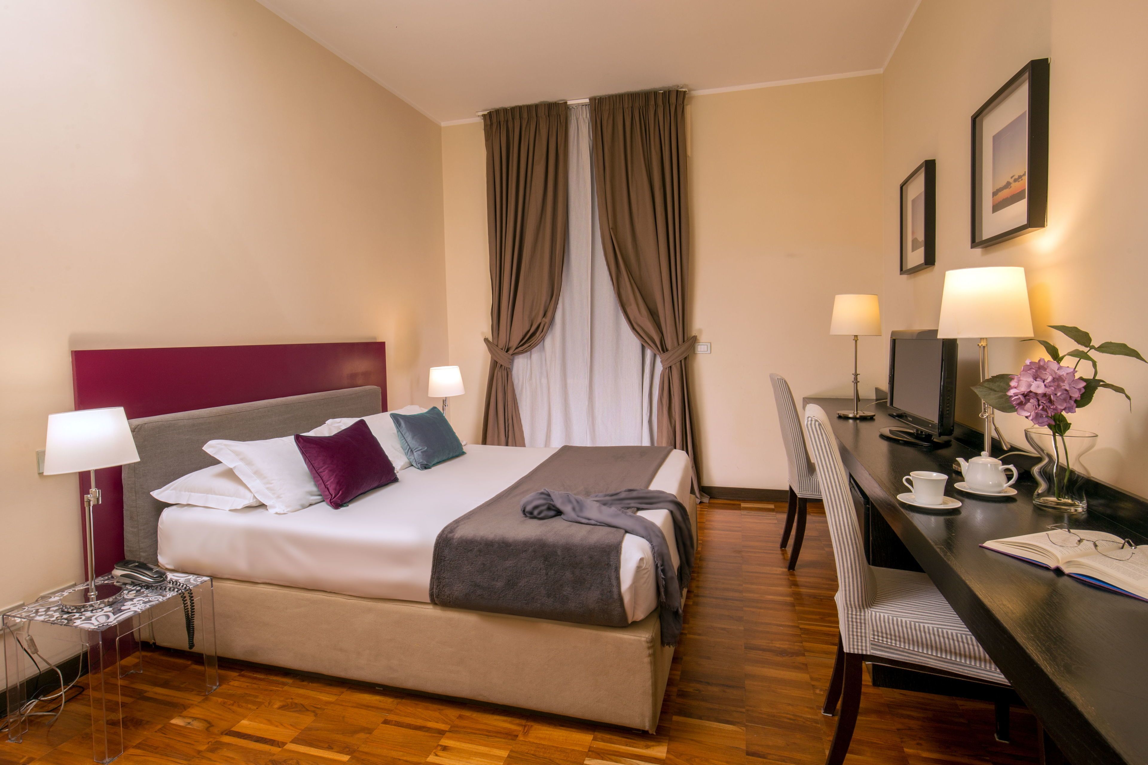 Ein Hotelzimmer mit Bett, Schreibtisch, Stuhl und einem kleinen Tisch mit Lampe und Blumenvase.