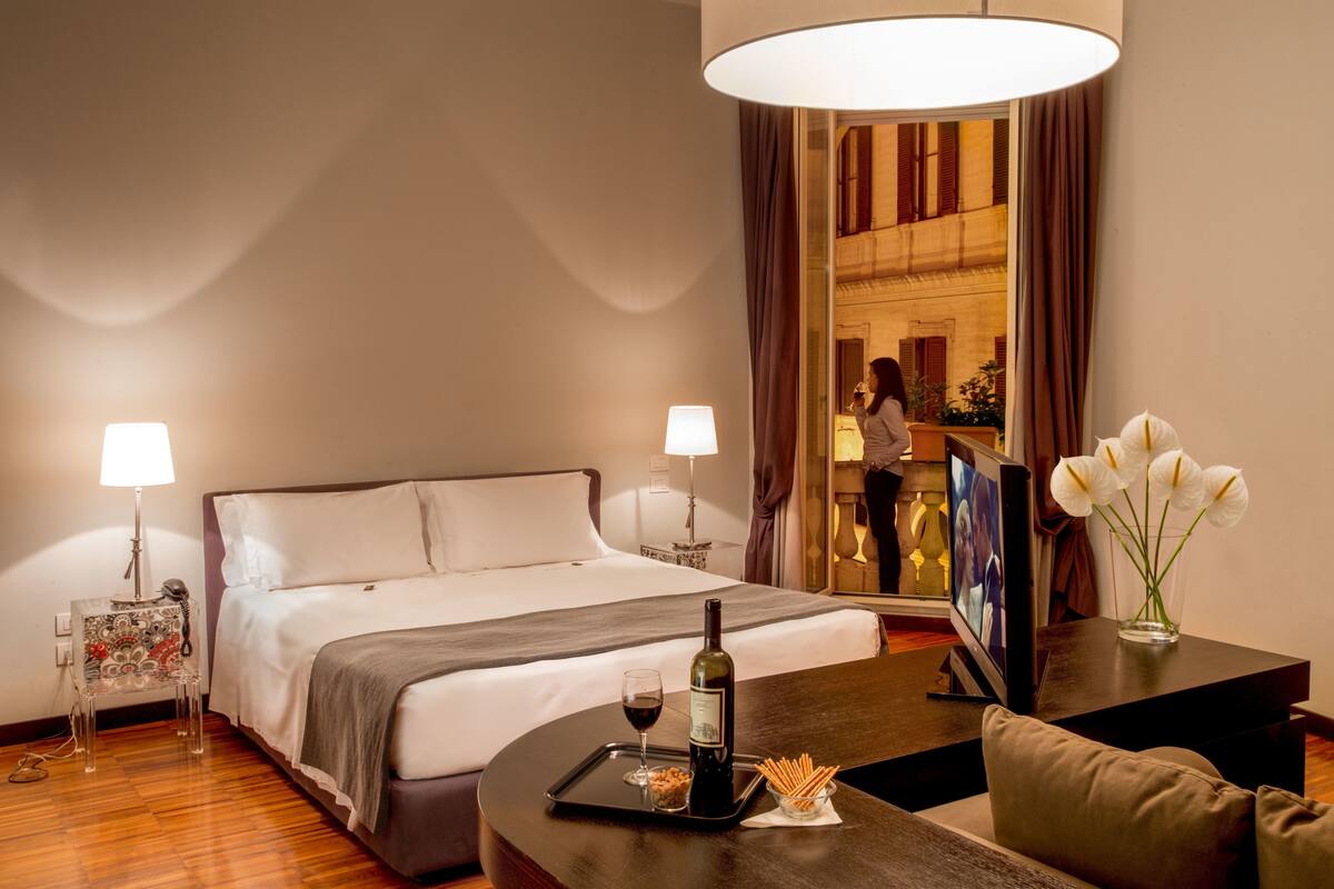 A hotel room with a bed, bedside lamps, a TV, a dining table with a bottle of wine, and a vase with flowers.