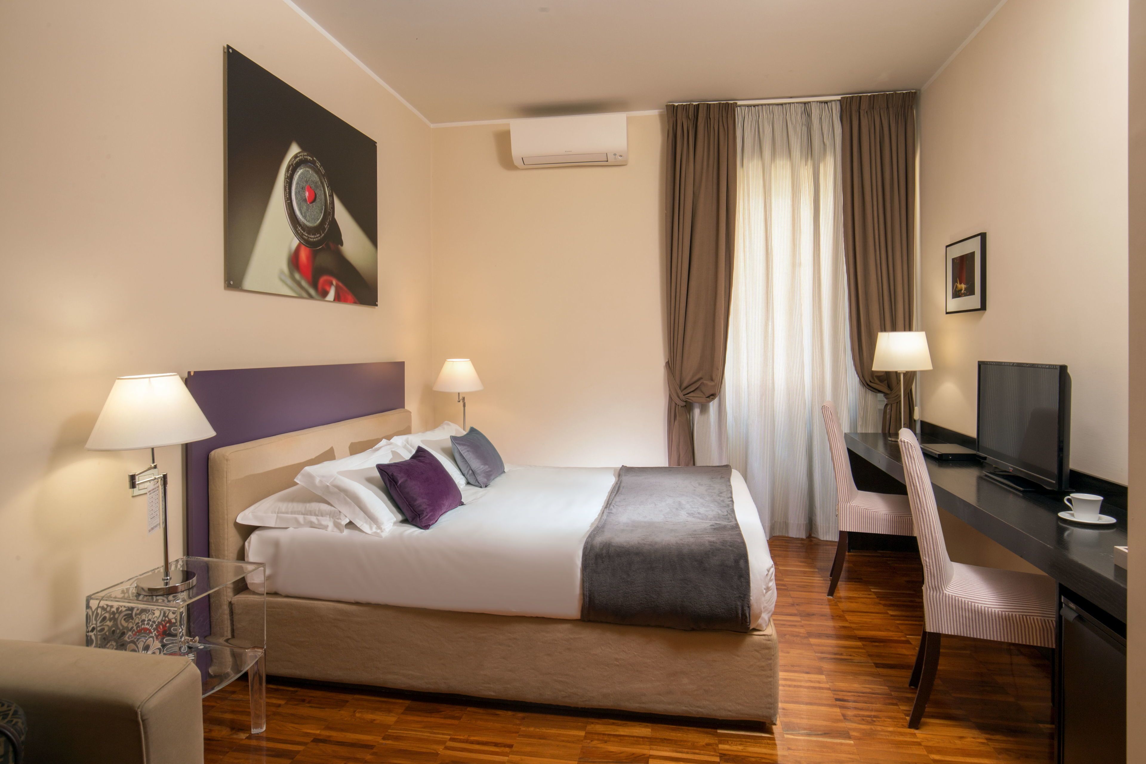 Una habitación de hotel con una cama grande, dos lámparas de noche, un escritorio con computadora, una silla y una ventana con cortinas.
