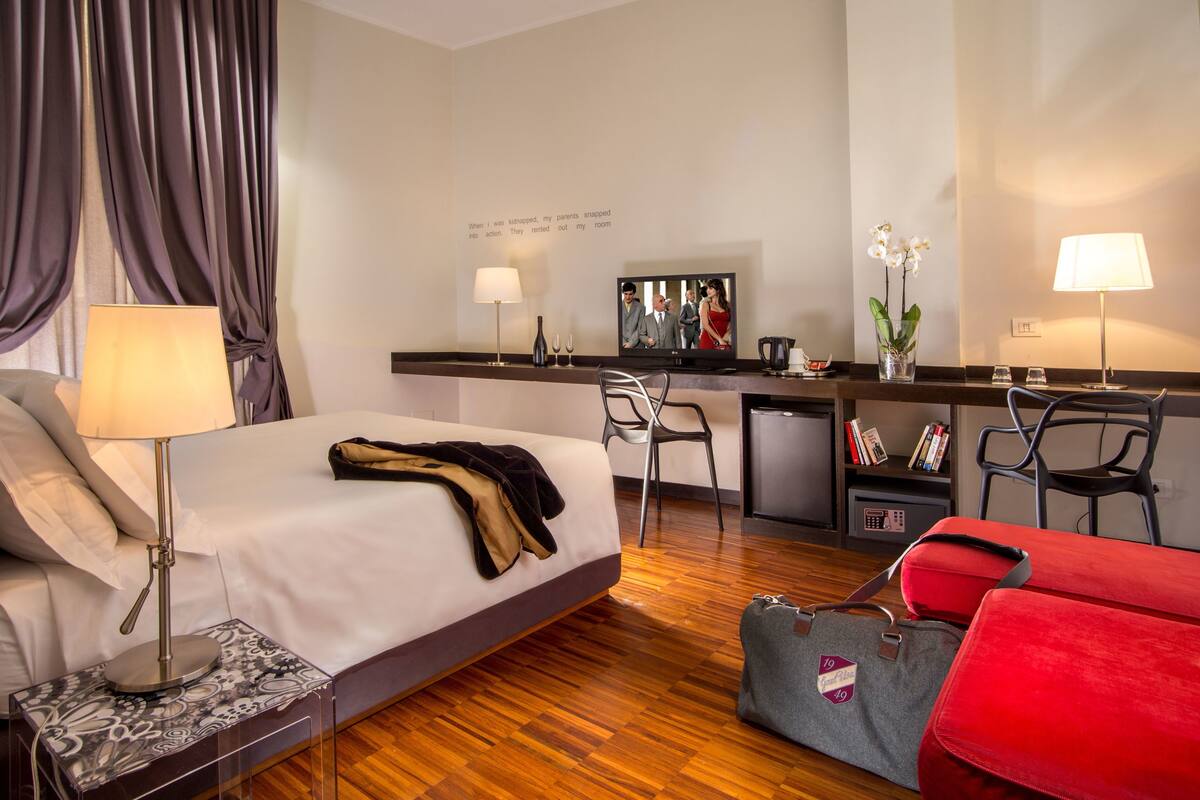 A hotel room with a bed, desk, chairs, a small kitchenette, and a bookshelf.
