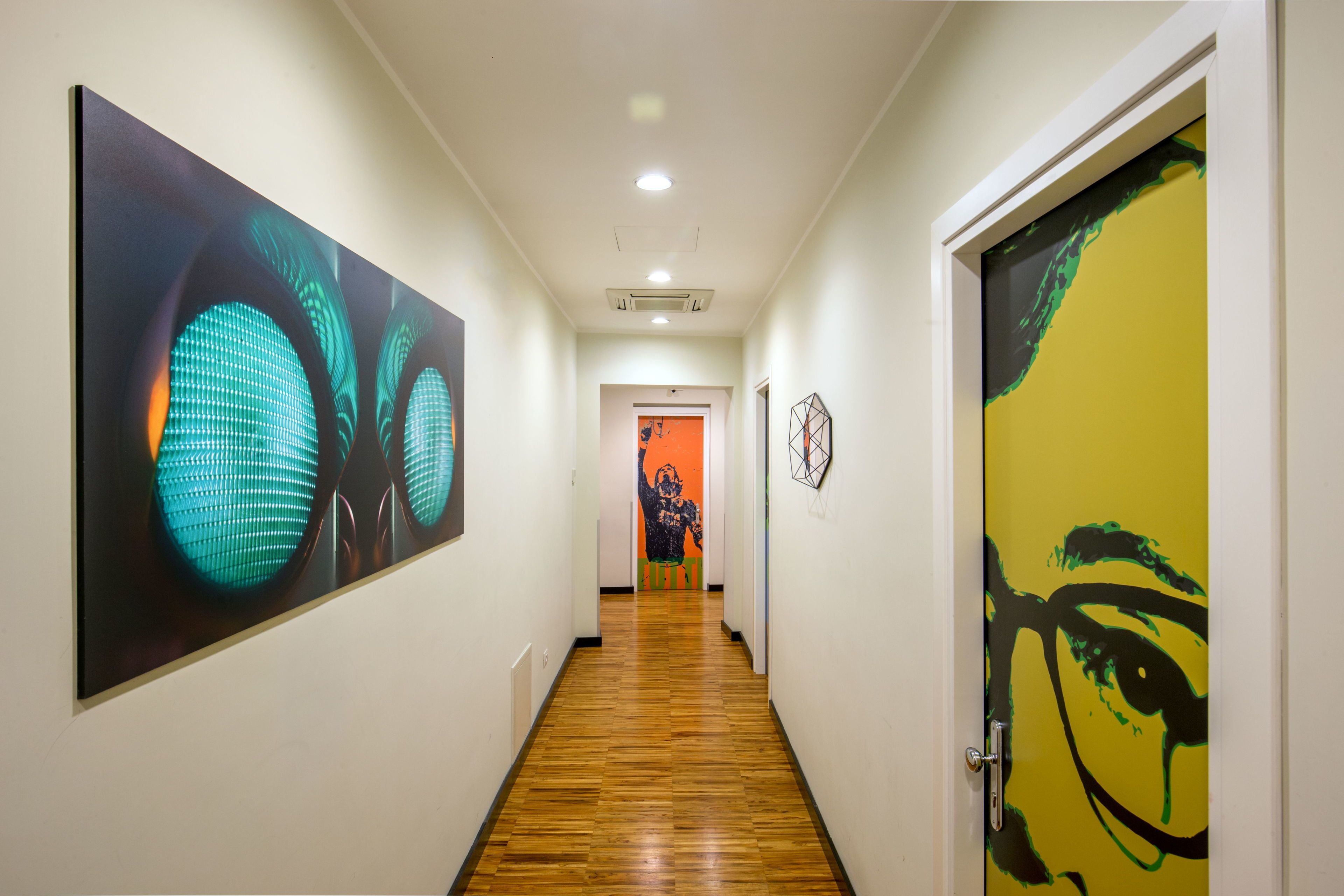 A hallway with wooden flooring, a door with a yellow and black design, and a large abstract painting on the left wall.