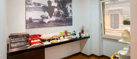 Una barra de desayuno con tostadora, fruta y pasteles, una foto en la pared y una ventana con vistas a la calle.