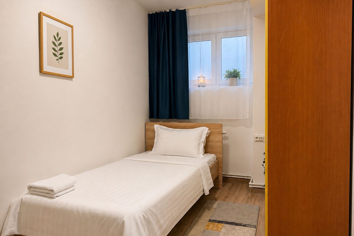A single bed with white bedding, a bedside table with a lamp, a window with curtains, and a framed picture on the wall.