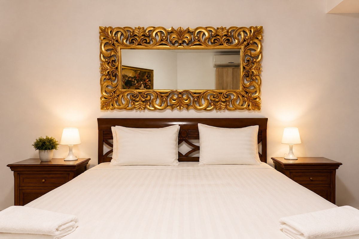 A hotel room with a large ornate mirror above the bed, two bedside lamps, and a neatly made bed with white linens.