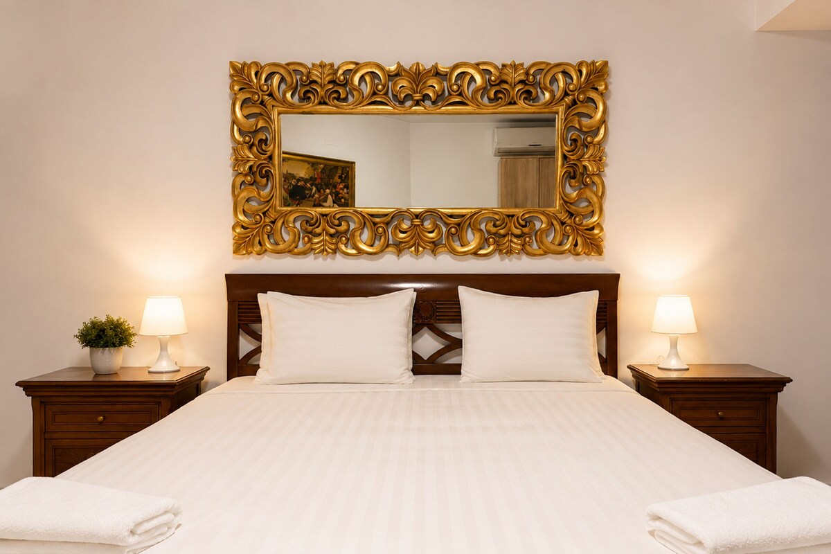 A hotel room with a large ornate mirror above the bed, two bedside lamps, and a neatly made bed with white linens.