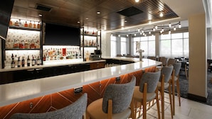 A modern bar area with a long counter, bar stools, and a variety of bottles. - Hilton Garden Inn Melville (Plainview)