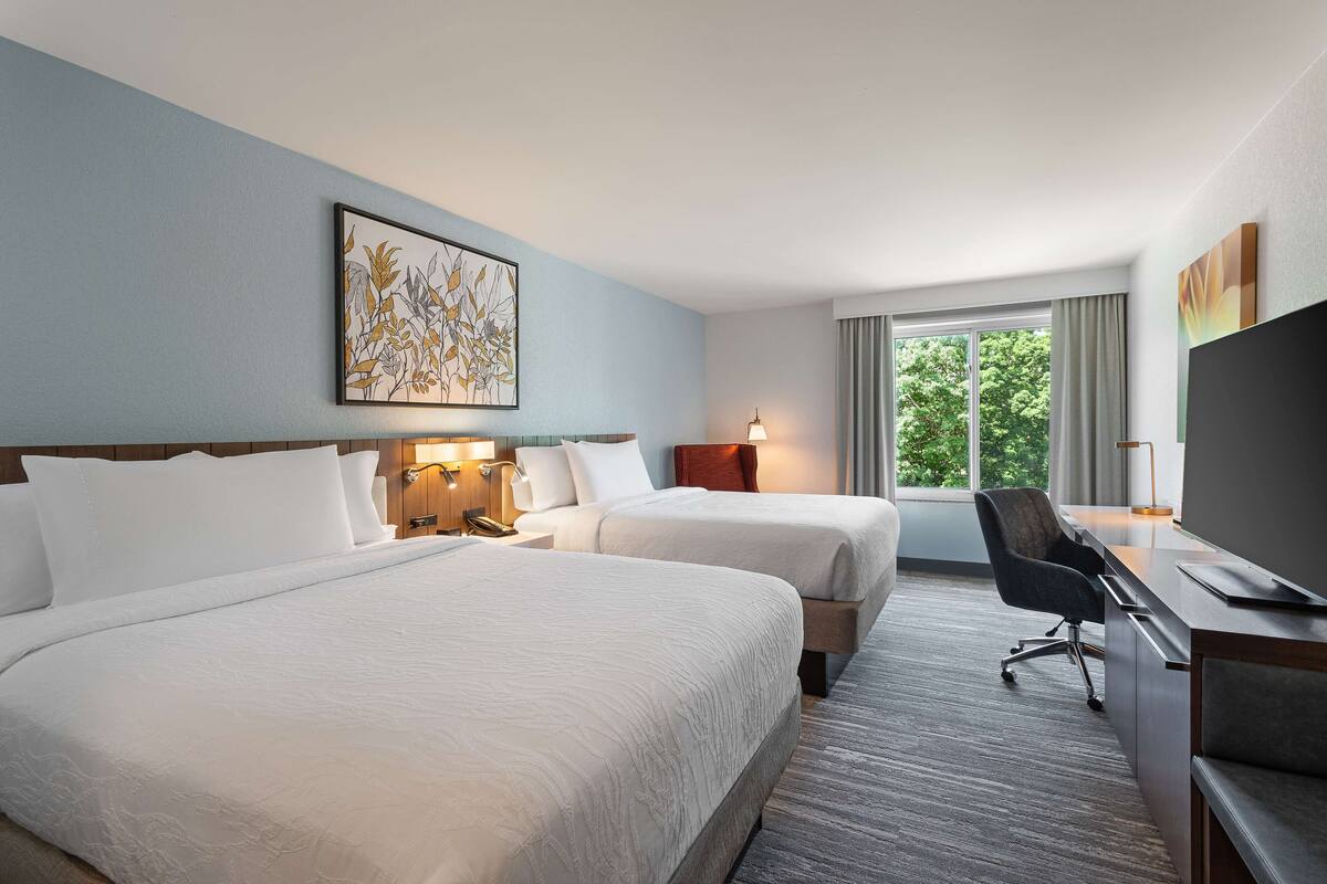 A hotel room with two beds, a desk, a chair, and a large window with a view of trees.