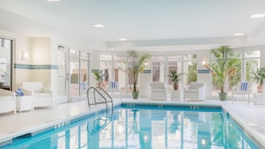 A modern indoor pool area with clear glass doors, white lounge chairs, and potted plants. - Hilton Garden Inn Melville (Plainview)