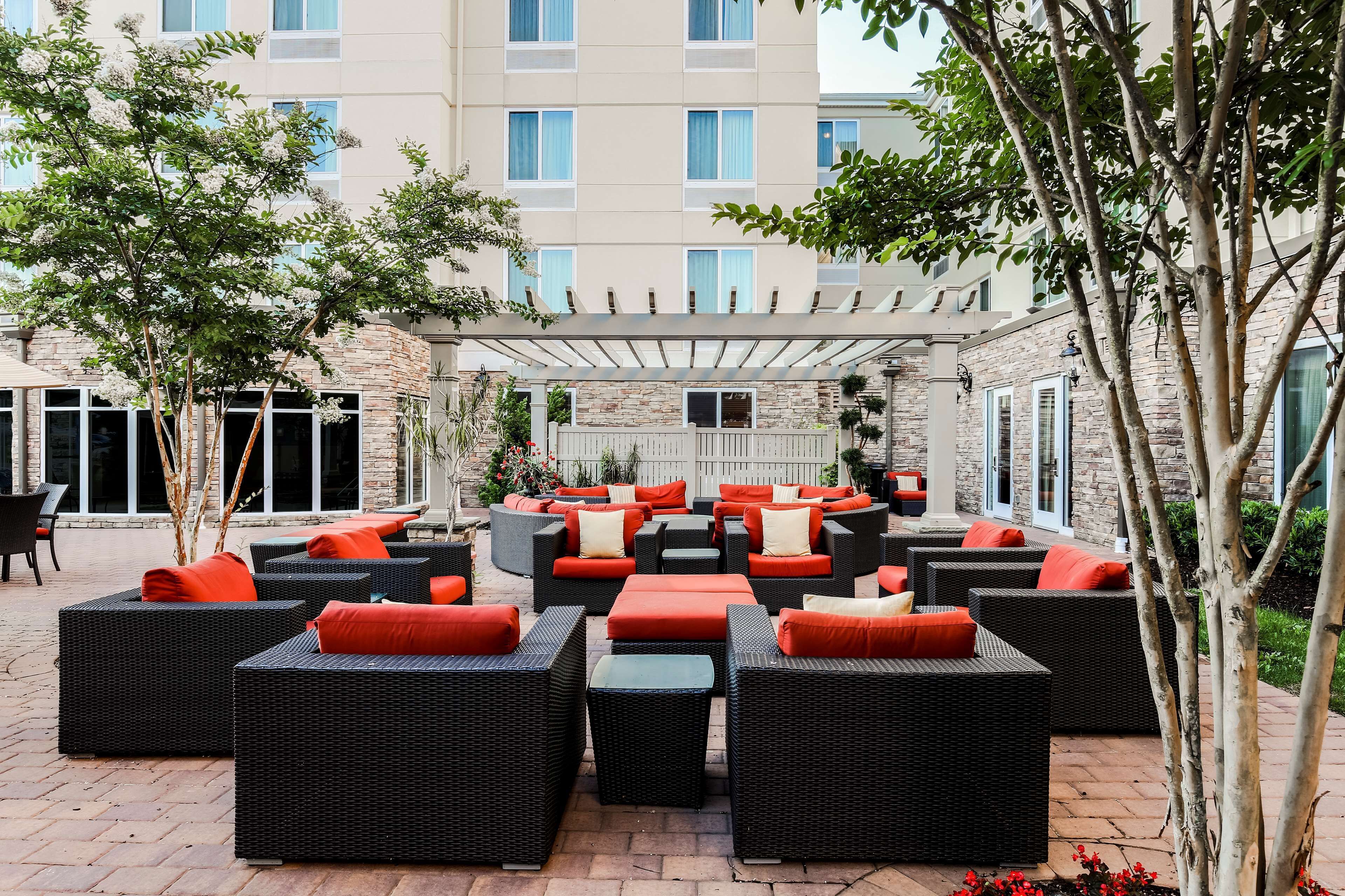 An outdoor seating area with wicker furniture and orange cushions, surrounded by trees and a building in the background.
