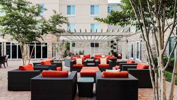 An outdoor seating area with wicker furniture and orange cushions, surrounded by trees and a building in the background.