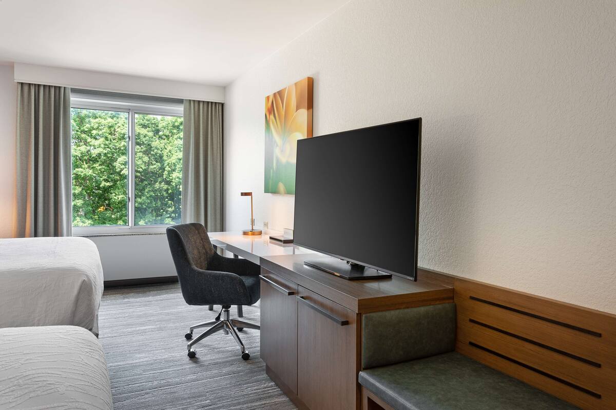 A hotel room with a bed, a desk with a flat-screen TV, a chair, and a window with curtains.