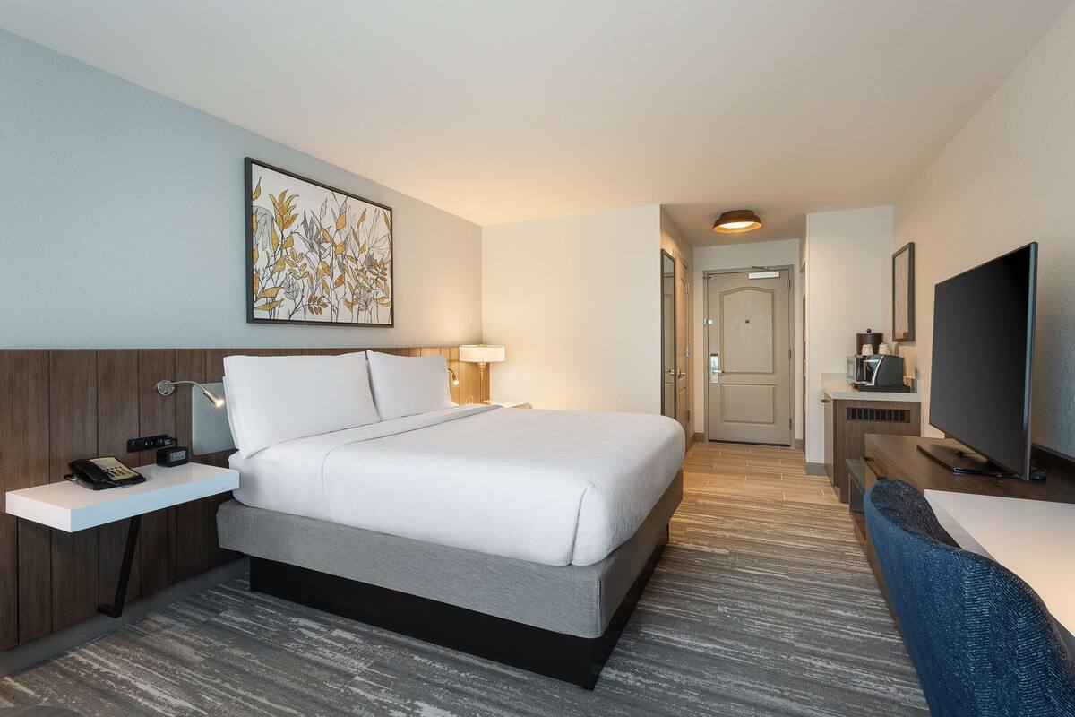 A hotel room with a bed, bedside table with a phone, a lamp, a TV, and a painting on the wall.