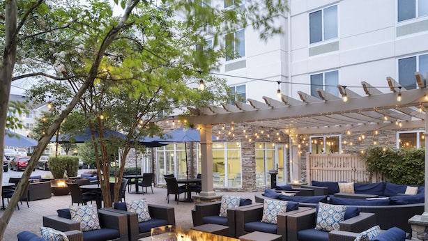 An outdoor patio area with wicker furniture, a central fire feature, and string lights. - Hilton Garden Inn Melville (Plainview)