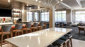 A modern hotel lobby with a bar area, seating arrangements, and large windows providing natural light. - Hilton Garden Inn Melville (Plainview)