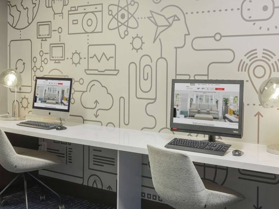 A modern office space with two desks, each with a computer monitor, keyboard, and mouse. There are two office chairs and two desk lamps. The wall features a circuit-like design with various icons.