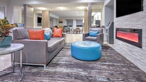 A modern hotel lobby with a grey sofa, blue ottoman, and a fireplace. - La Quinta Inn & Suites by Wyndham Alvarado (Alvarado)