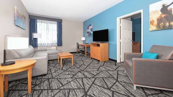 A hotel room with a grey sofa, a grey armchair, a wooden side table, a TV on a wooden stand, and a patterned carpet.