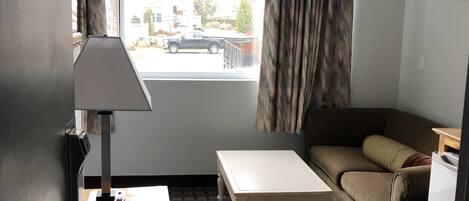 A small, furnished room with a sofa, a table, a lamp, and a window with curtains.