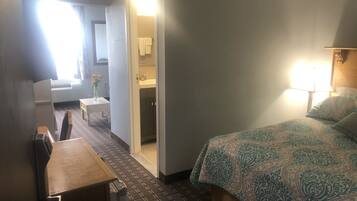 A hotel room with a bed, a desk, a chair, a small table, a bathroom with a sink and mirror, and a window with curtains.