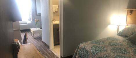 A hotel room with a bed, a desk, a chair, a small table, a bathroom with a sink and mirror, and a window with curtains.