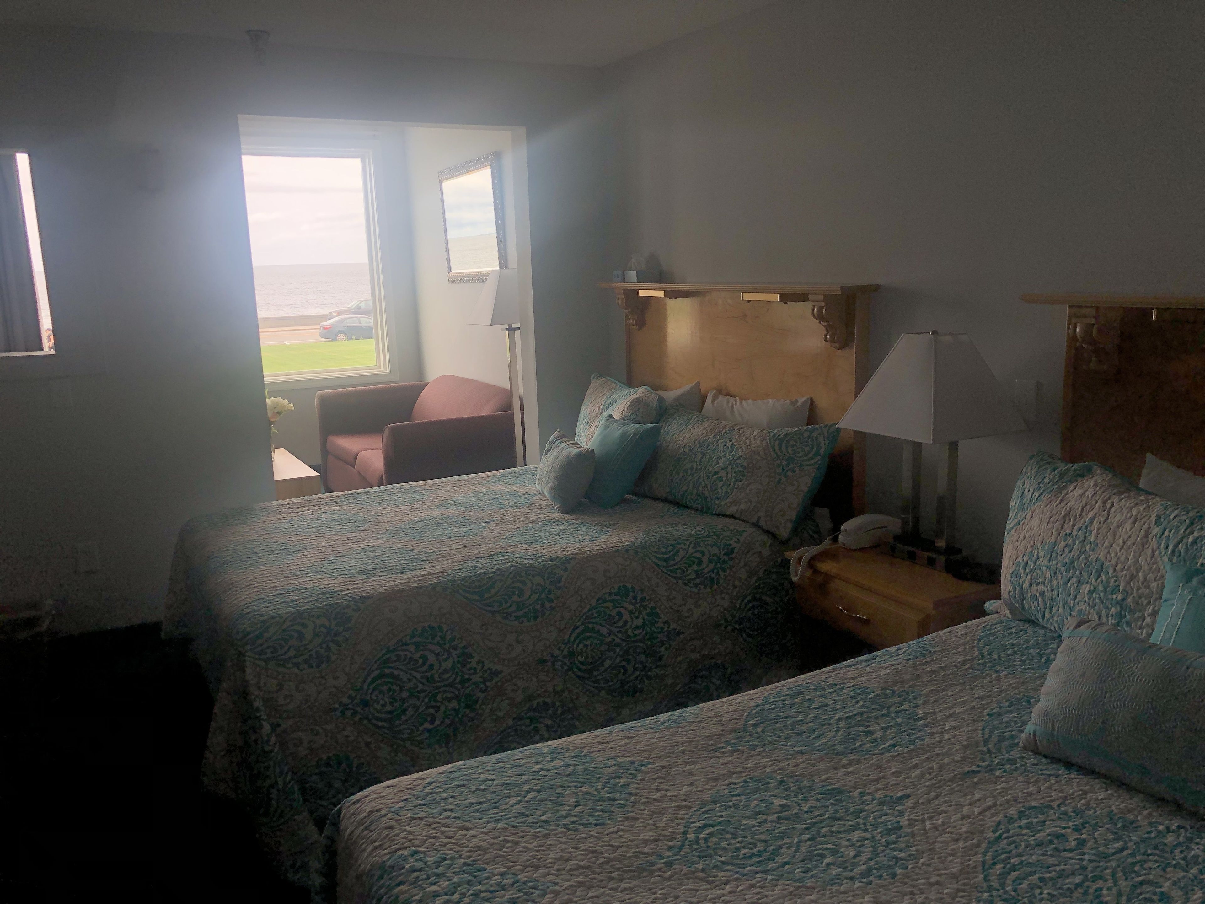 A hotel room with two beds, a nightstand, a lamp, a chair, and a view of the outdoors.