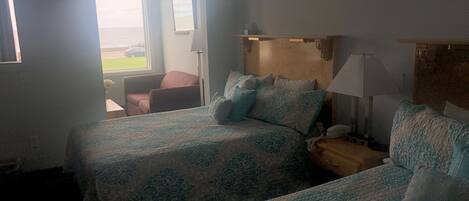 A hotel room with two beds, a nightstand, a lamp, a chair, and a view of the outdoors.