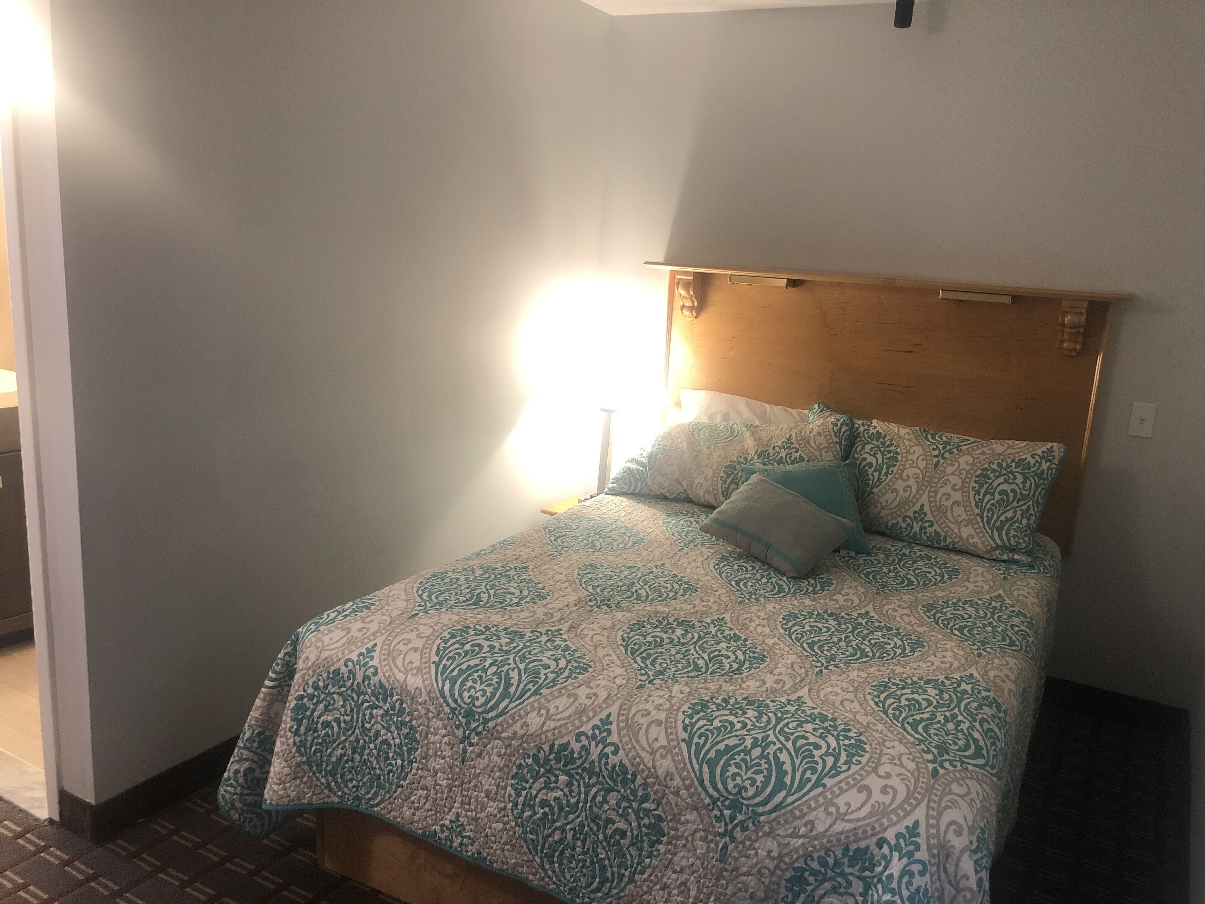 A bed with a patterned bedspread and pillows, a wooden headboard, and a bedside lamp.