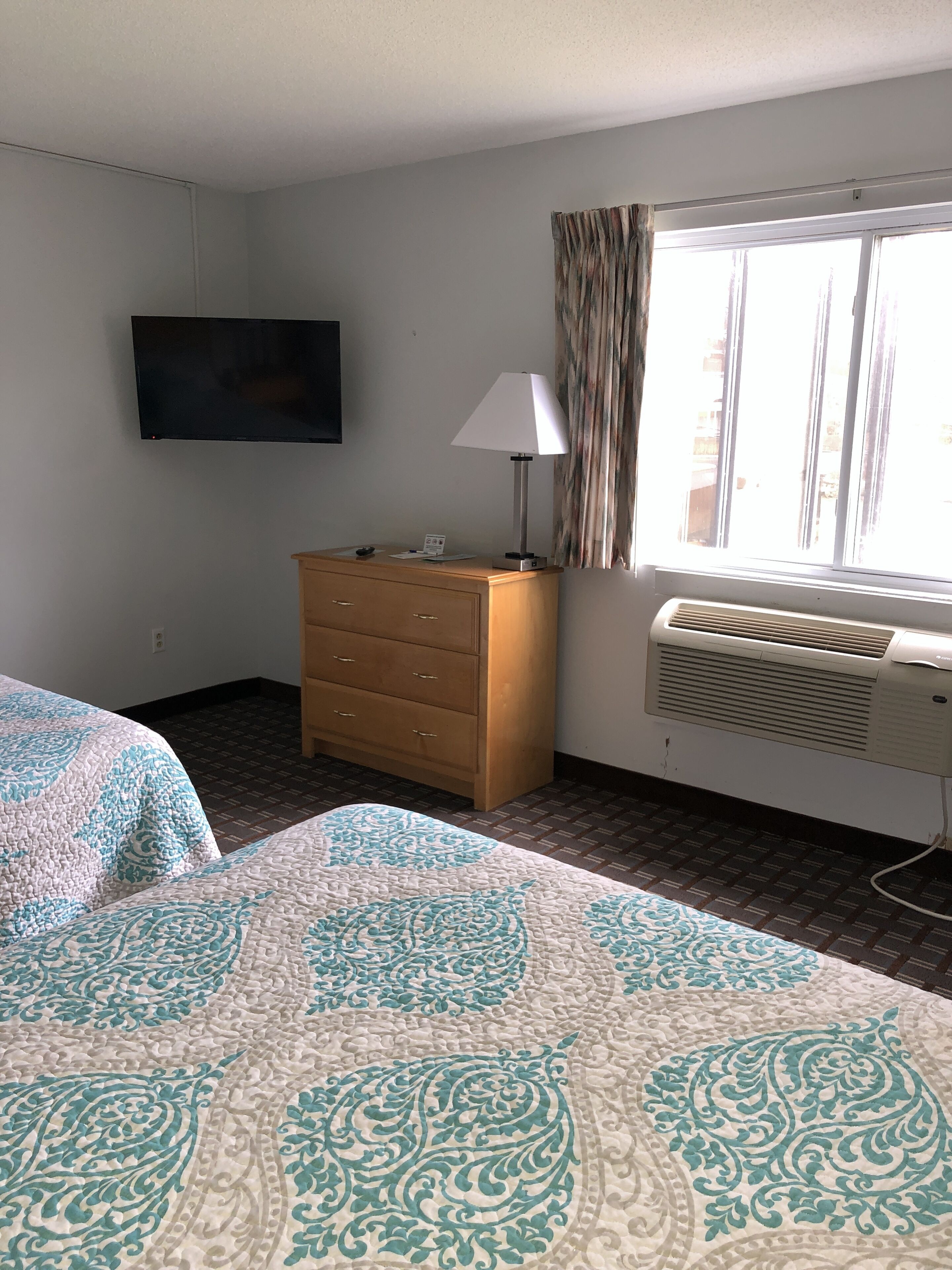 A hotel room with two beds, a wooden dresser, a flat-screen TV, a lamp, and an air conditioning unit.