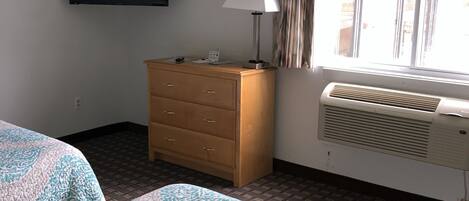 A hotel room with two beds, a wooden dresser, a flat-screen TV, a lamp, and an air conditioning unit.