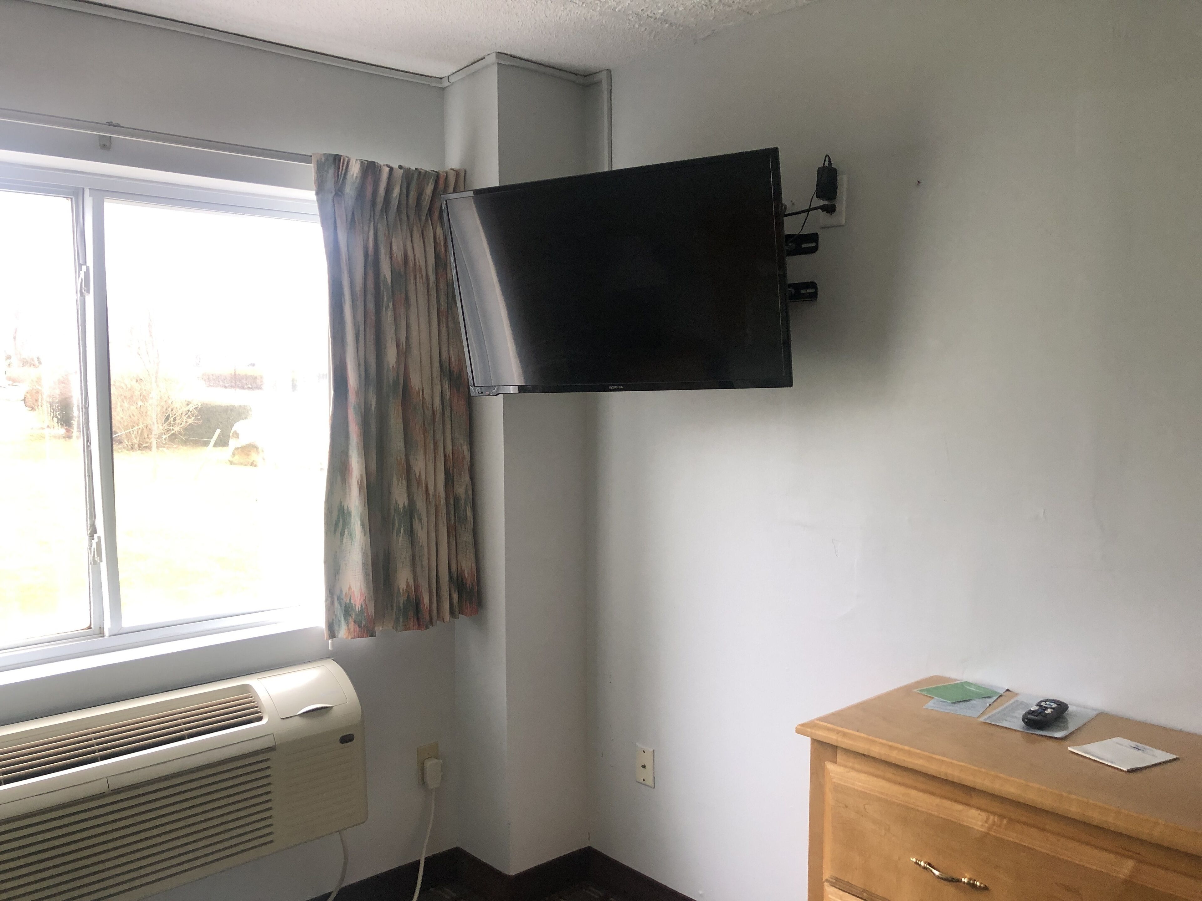 A room with a window, an air conditioner, a flat-screen TV mounted on the wall, and a wooden dresser with a few items on top.