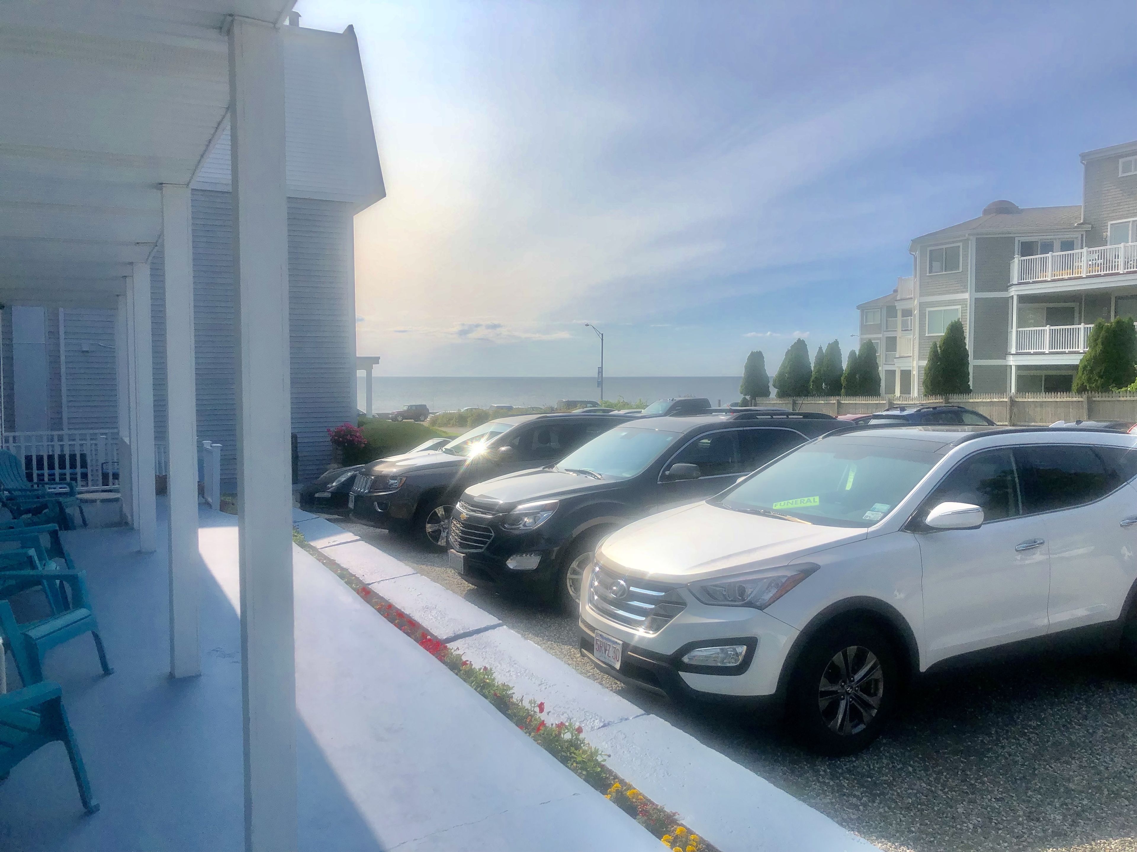 Un parking avec plusieurs voitures, un bâtiment à la façade blanche et une vue sur l’océan.